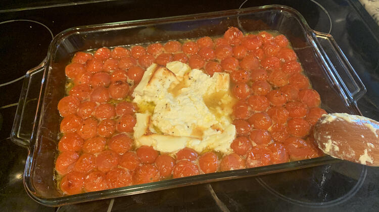 Image shows the glass casserole dish filled with deflated tomatoes in liquid and a slightly misshapen block of feta in the center. You can see the spoon section of the wooden utensil as it rests over the corner of the casserole dish. 