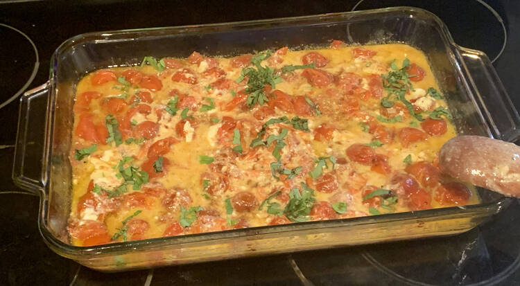 Image shows a yellow-ish liquid in a glass casserole dish filled with round red tomatoes, chunks of white feta, dark splotches of balsamic vinegar, and slivered green basil. The spoon, once again, is leaned up over the corner of the dish. 