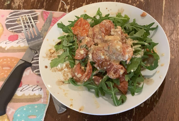 Image shows a plate that appears to hold a salad as the single piece of toast is hidden underneath. It shows the tomato and feta mixture on top of a pile of arugula. Beside the plate sits a donut decorated napkin underneath a dirtied fork and steak knife. 