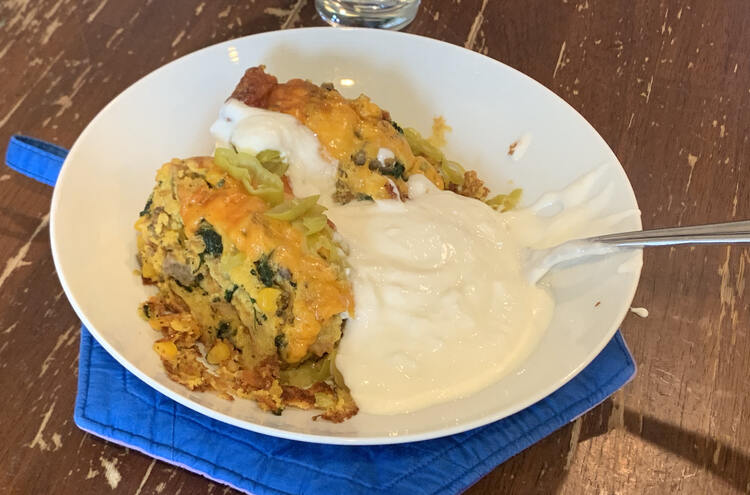 Image shows a white bowl with the orange and tan dish with white yogurt and sliced pepperoncini. There's a spoon hiding in the large pool of yogurt surrounding the dish. Under it sits a blue potholder and the table. 