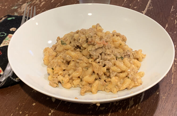 Image shows a mound of elbow macaroni coated in orange in the center of a white bowl with a pizza themed fabric napkin and fork off to the left side.