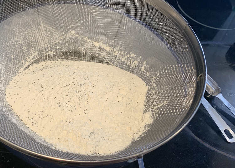 Image shows a metal strainer over a white (inner) bowl with white powder with black specks in it. There are two measuring spoons in the background. 