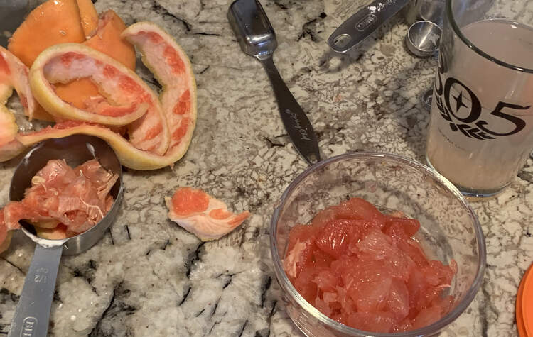 Image shows a glass container with grapefruit pulp. Beside it (bottom left, up, and around) is a measuring cup with clear grapefruit edges, the cut peels, a couple measuring devices, and a pink hued water glass. 