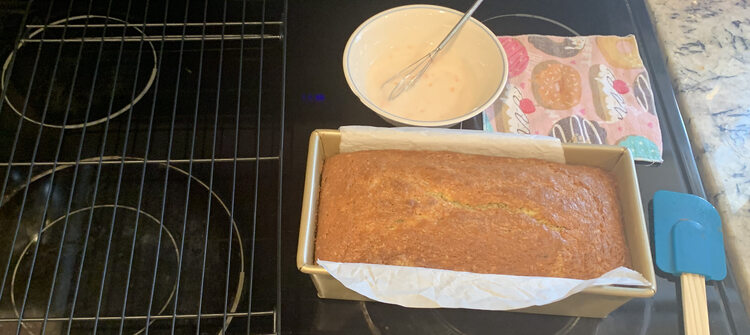 Image shows a baked poppy seed loaf siting in a loaf pan with parchment paper sticking out from either long side. Beside it sits a blue spatula, a napkin, a bowl with icing and a small whisk, and a wire cooling rack. 