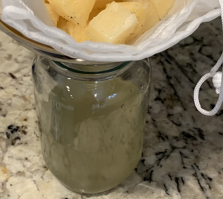 Image shows a mesh bag holding black flecked pineapple on a funnel over a yellow liquid filled clear mason jar. 