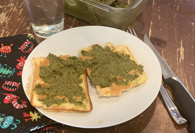 Image shows two waffles with green mixture on top on a white plate. Beside it sits a fork and knife on one side and a monster fabric napkin on the other. Behind it sits a water glass and a glass container of more pistachio pistou.