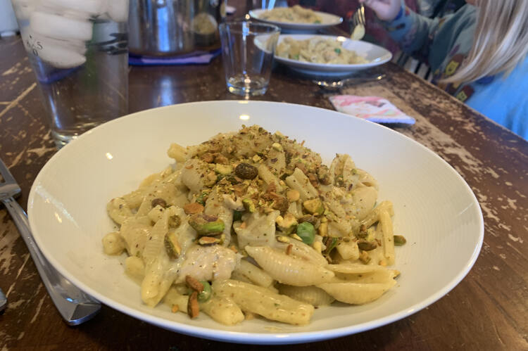 Image shows a white bowl with a mound of green flecked pasta topped with crushed nuts. In the background is the pot and the girls' plates.  