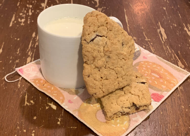 Image shows a white mug filled with milk on a cupcake decorated napkin on the kitchen table. Beside it sits one cookie, lying flat, and another leaned up vertically on the first cookie and leaning against the mug. 