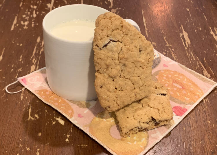 Image shows a white mug filled with milk on a cupcake decorated napkin on the kitchen table. Beside it sits one cookie, lying flat, and another leaned up vertically on the first cookie and leaning against the mug.