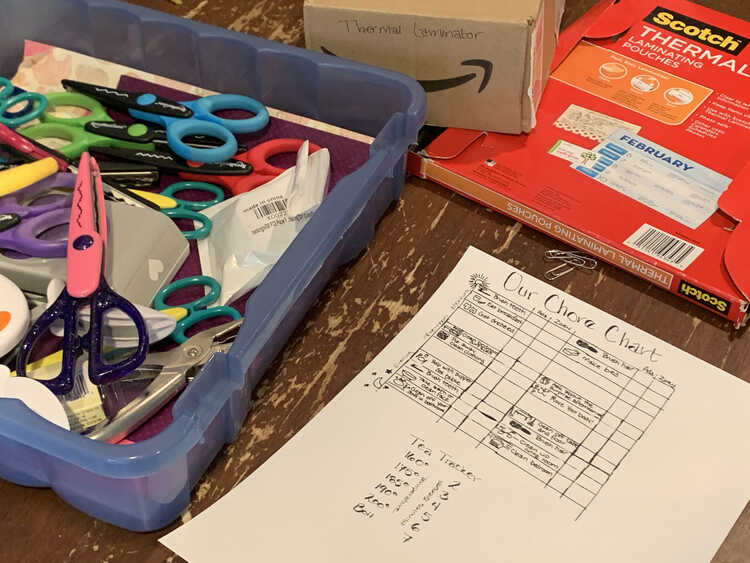 Image shows the sheet of paper with a chore chart above the tea tracker. Beside it is a blue scrapbook paper container filled with paper, decorative scissors, and hole punches. Behind it sits a thermal laminator and package of laminating pouches. 