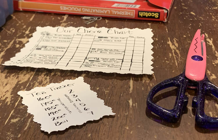 Image shows the Tea Tracker sitting on a small rectangle of paper with the edge scalloped. In the background sits the similarly trimmed chore chart, matching scissors, and the laminating pouches. 