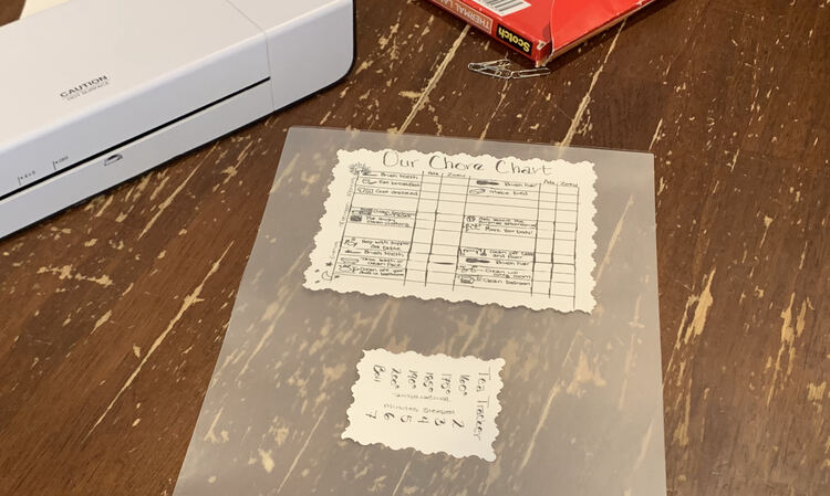 Image shows most of the laminator pouch with the larger chore chart closer to the top and the Tea Tracker turned on it's side closer to the bottom of the photo. In the background sits the thermal laminator and the package of laminating pouches. 