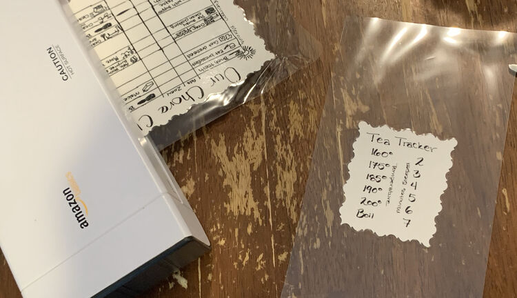 Image shows the Tea Tracker laminated within a tall sheet of lamination. In the background the chore chart is going into the thermal laminator sideways to remove the wrinkles on the top left side. 
