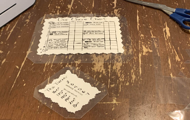 Image shows the laminated and trimmed Tea Tracker (in the front), Chore Chart (further to the back, scissors, laminator, and bits of scrap. 