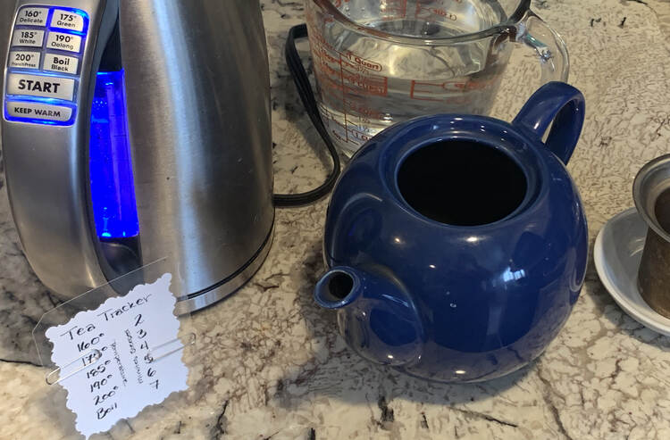 Image shows an electric kettle heating up to 175° while the tea tracker below has paperclips highlighting the 175° temperature and 5 minutes steeped. Beside it sits a blue teapot with the infuser beside it on a white mini saucer. Behind everything sits a four cup glass jug with water in it.