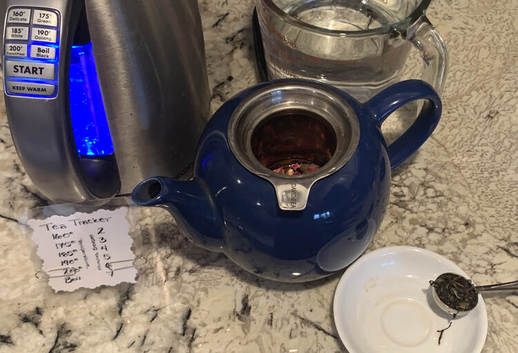 Image shows a scoop of black tea on the white saucer, a blue tea pot with rose petals showing in the infuser, my tea tracker with the paper clips set to 200° and 6 minutes, the kettle set to boil, and a glass jug full of water. 