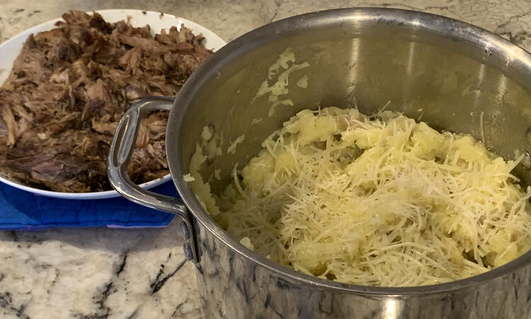 Image shows a metal pot, to the right, partly filled with white potatoes topped with shredded parmesan cheese. In the background, to the left, sits a shredded pork roast in a white bowl on a sewn trivet. 