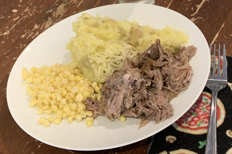 Image shows a white plate next to a fork and pizza themed napkin. On the plate sits three piles all touching. The top is the potatoes with lattice work of parmesan cheese on the side closest to us. Beside it is a pile of yellow corn and shredded meat. 