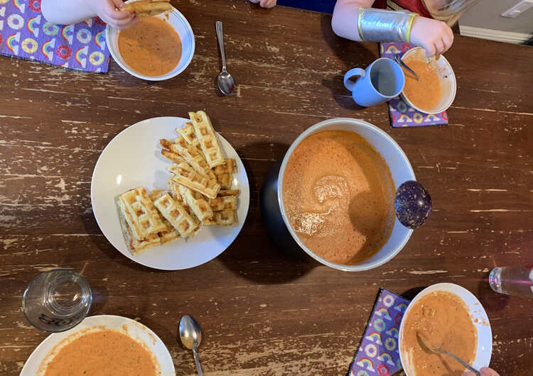 Image is taken from above showing our kitchen table with the liner of blended tomato soup next to a plate of cheesy waffle dippers. Four bowls are around it with waffles being dipped and eaten above the two smaller kid ones. 