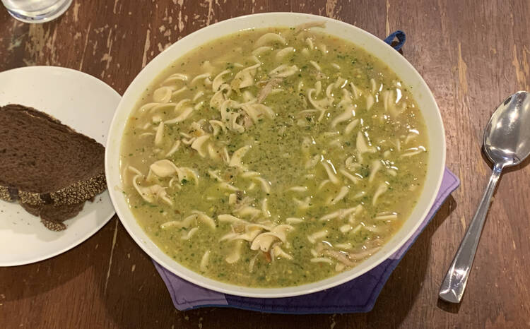 Image is taken from above showing a bowl filled with green flecked soup and egg noodles. To the left sits a saucer with pumpernickel toast and to the right a large spoon. 