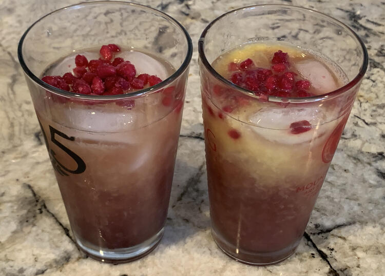 Image shows two clear glass cups side by side with ice and red-ish liquid and pomegranate seeds on top. 