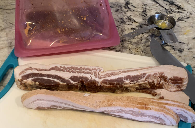 Image shows two stacks of raw bacon on a plastic cutting board. Beside it sits a sharp knife and behind it a pink Stasher bag on its side with the ingredients mixed within. 