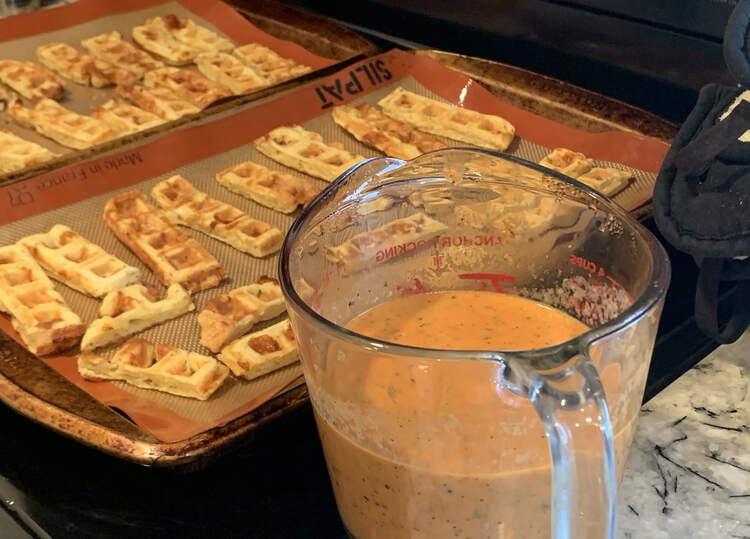 Image shows two lined cookie sheets covered in freshly toasted waffle dippers with a jug of reheated and pourable soup ready to go. 