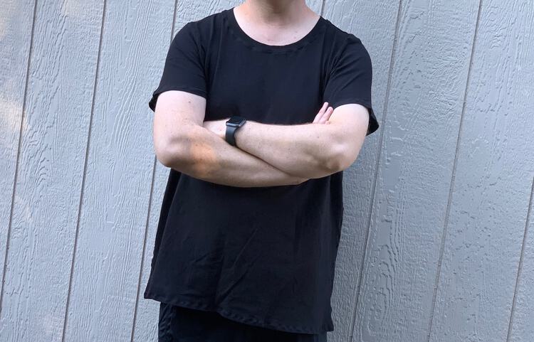 Image shows the torso of a white man wearing a black tee with his arms crossed in front of him. He stands in front of a white wall making his shirt pop a bit. Image is taken from his neck to right below the tee. 