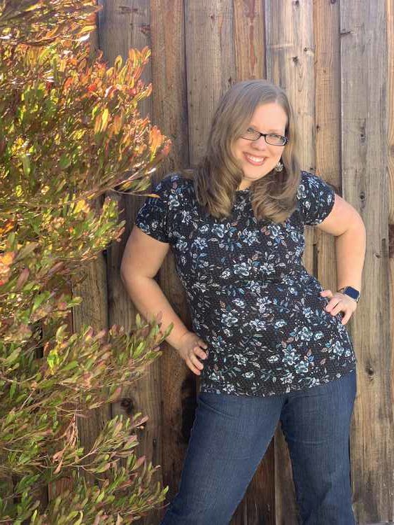 Photo of my next to a tree with a fence behind. My hands are on my hips, hip jutted out, and hair curling in below my shoulders.