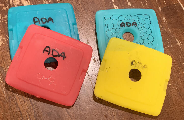 Image shows four ice packs on the table. The two on the left show white remnants from where the original design was and are recently marked with "Ada" in bold black paint. On the right is the newly flowered pack, behind, and a scrapped off yellow ice pack, in the front, with the word "Ada" vaguely legible.