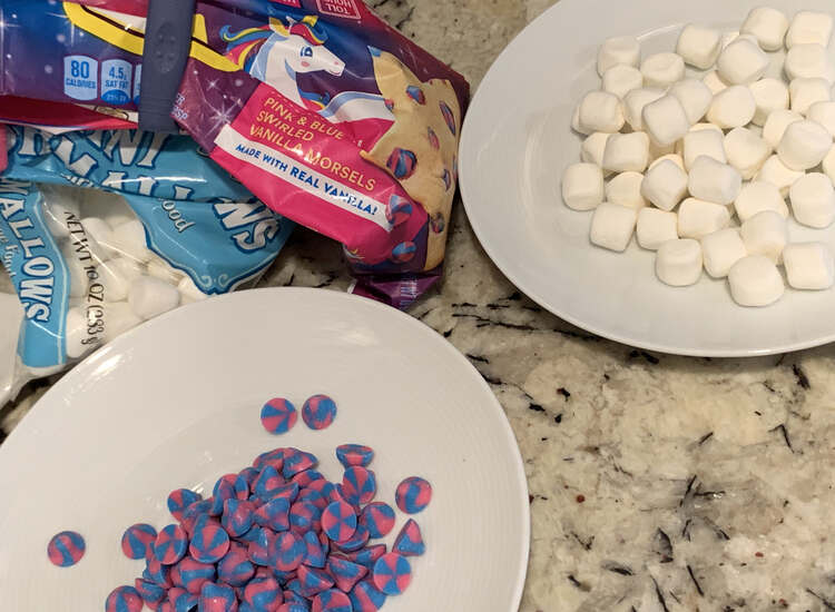 Image shows two white saucers one with white marshmallows, to the right, and one with pink and blue swirled chocolate chips, to the left. In the background you can see the bags of the leftover ones. 