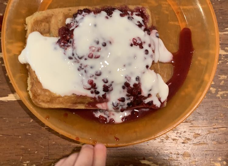 Image shows a waffle on an orange plastic plate. IT's topped with a red bumpy sauce with some red liquid along the bottom right edge. Overtop of most of it is a white sauce. 