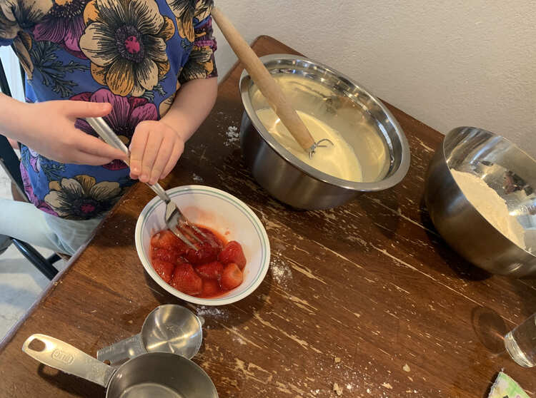 Zoey uses a fork to squish whole strawberries in a small bowl on the table. Behind it sits two large bowls one with wet ingredients and one with dry. 