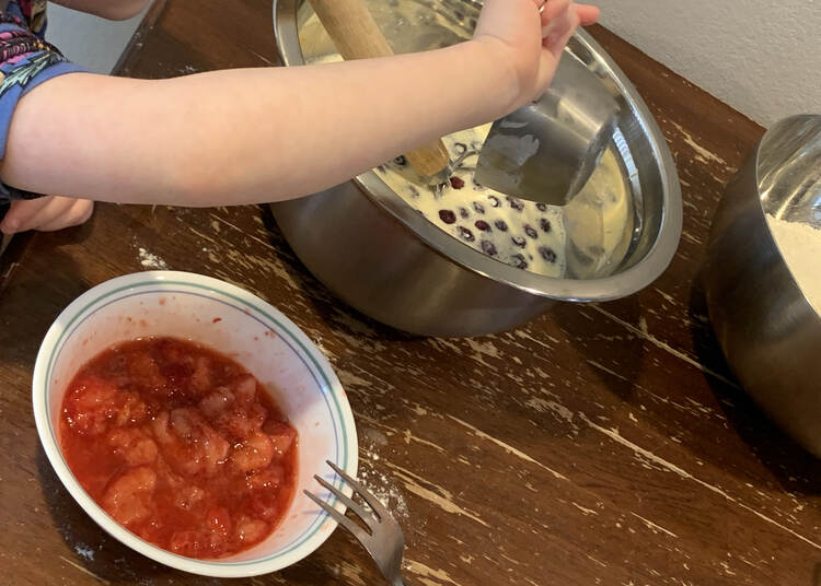 Zoey dumps a measuring cup full of blueberries into the wet ingredients of the pancake batter. In front of it sits a small bowl of crushed thawed strawberries. Off to the side in the back sits a bowl of dried ingredients. 