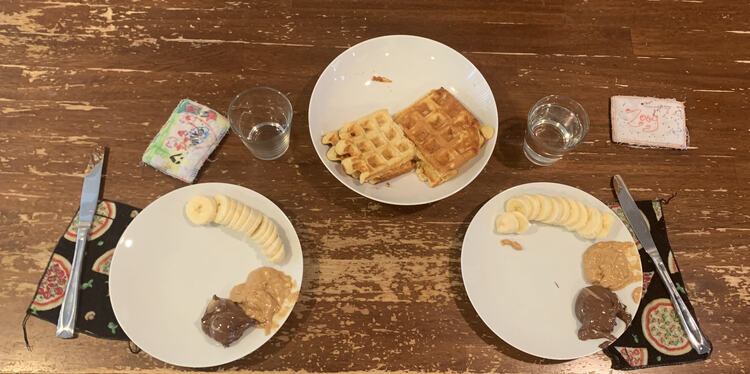Image shows three plates laid out. The two closes to the bottom each have a banana sliced up, a dollop of peanut butter, and a dollop of Nutella. They have a knife, napkin, glass, and nametag nearby. Above and in the center sits another plate with four waffles on it waiting to be eaten. 