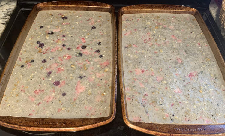 Image shows two similar cookie sheets of pancakes side by side. The right one has blue tinged batter with pink strawberry chunks through it. The left one looks the same with more blue circles of blueberries in it. 