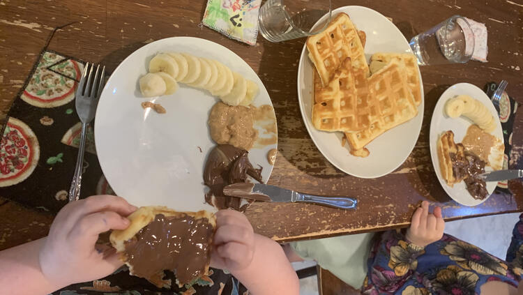 Image shows more waffles on the center plate and this time the girls are sitting at their plates eating or decorating their waffles. 