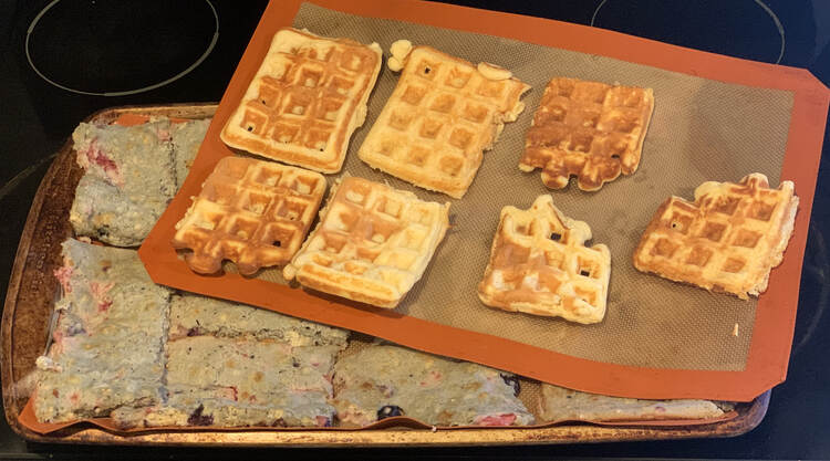Image shows A cookie sheets with a Silpat filled with blue tinged pancakes. On top, at an angle, sits another Silpat with seven waffles lined up on it. 