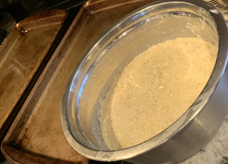 Image shows a bowl half full of pancake batter with two greased cookie sheets sitting beside each other. 