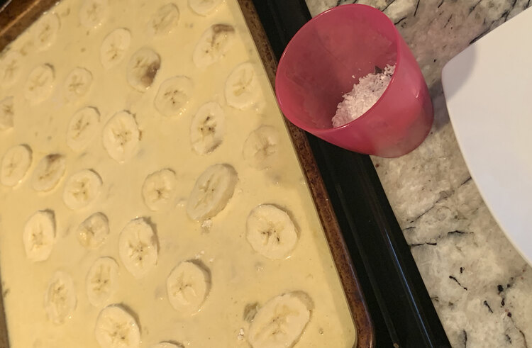 Image shows a sheet pan of pre-cooked pancake dough with sliced bananas lined up in it. To the side is an empty saucer and a pink plastic cup with coconut and chocolate chips inside. 