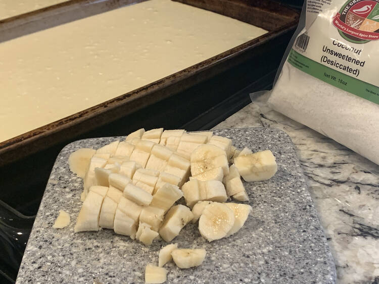 Image shows two cookie sheets with pancake batter in the background. In the front sits a marbled cutting board with bits of banana and beside that sits a bag of unsweetened coconut flakes from The Great American Spice Company.