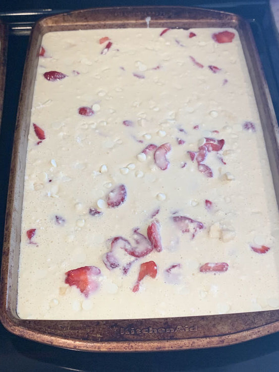 Image shows uncooked pancake batter in a sheet pan with pink strawberries showing through the dough and white chunks of sinking chocolate chips. 
