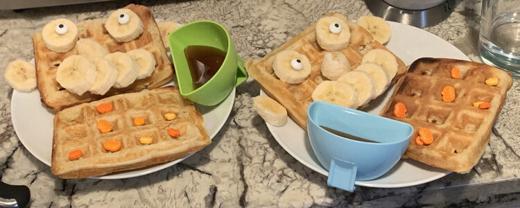 Image shows two saucers with two waffles each and a plastic dipper with syrup. One waffle on either plate holds pumpkin sprinkles while the other one looks like a person with banana ears, nose, mouth, and eyes. 