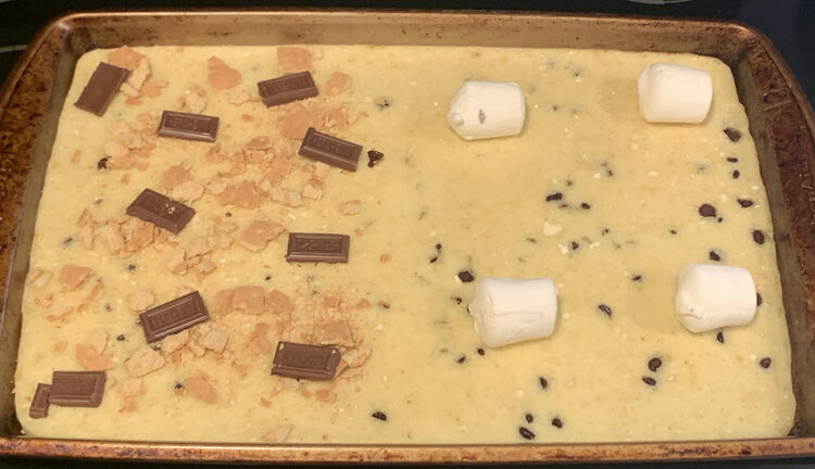 Image shows a sheet pan of baked pancake with the odd chocolate chip showing through from the bottom. The left side has rectangles of chocolate on it surrounded by graham crumbs. The right side has four evenly spaced large marshmallows with a hint of brown on one. 