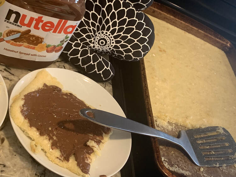 Image shows a cookie sheet with a strip of pancakes missing and a metal flipper on the right. On the left is a jar of Nutella, an oven mitt, and a play with a Nutella decorated square pancake on it. 