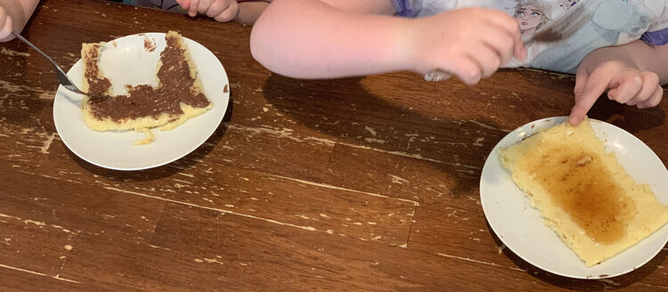Image shows two plates with the girls about or currently eating it. The one half devoured on the left is coated in Nutella while the one on the right is whole and holds a bowl of syrup in the center. 