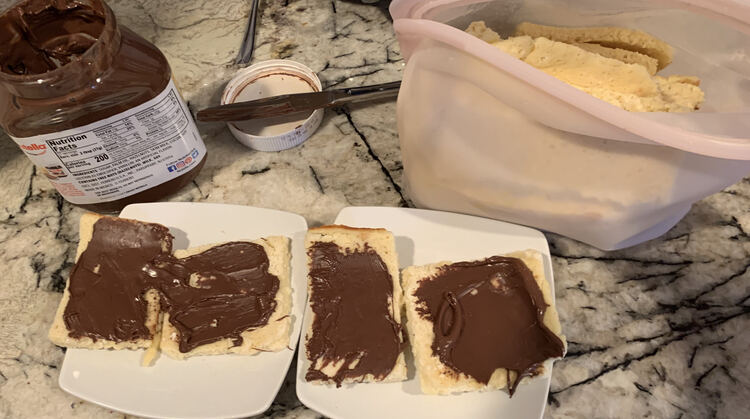 Image shows two square saucers, side by side, with two Nutella spread pancakes on top. A container of Nutella sits behind it along with a dirty knife and a full upright Stasher bag of more pancakes. 