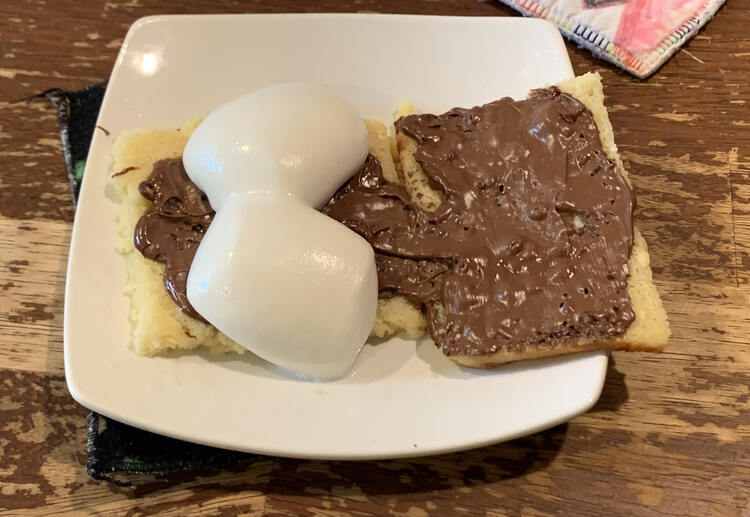 Image shows two heated pancakes with Nutella on both and two marshmallows on the left. 