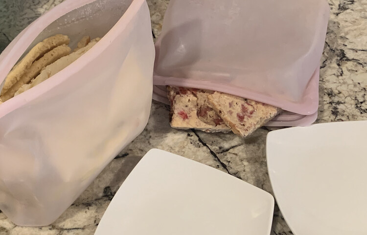 Image shows two Stasher bags popped open. The one on the left is upright and the one on the right is on it's side with some spilling out. In front of them are two white square saucers. 