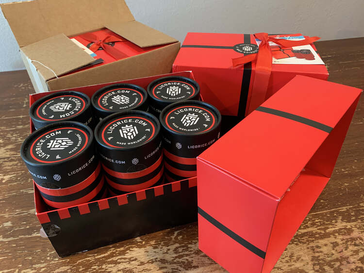 Image shows three sets of six licorice gift packs. The back box is still within a shipping box. To the right sits a closed box with the ribbon still wrapped around it. In the front sits an opened gift box showing the six licorice canisters. 